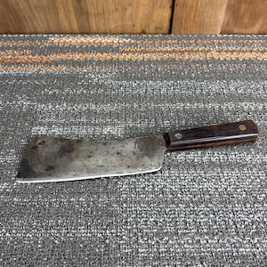 May include: A vintage cleaver with a dark wooden handle and a worn, rectangular blade. The blade shows signs of use and age, with a slightly curved cutting edge. A brass rivet secures the handle to the blade.