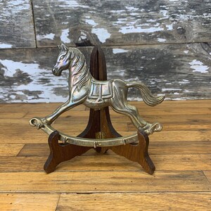 May include: A silver rocking horse figurine on a wooden stand. The horse is detailed with a saddle and bridle.