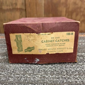 May include: A maroon cardboard box with a label that reads "DIE CAST CABINET CATCHES." The label also includes the text "BRIGHT CHROMIUM PLATED COMPLETE WITH SCREWS" and the manufacturer's name, "HIBBARD, SPENCER, BARTLETT & CO., EVANSTON, ILLINOIS."