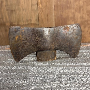 May include: A vintage double-bit axe head with a weathered, rusty appearance. The axe head is supported by a small, round wooden block. The metal has a dark, aged patina, suggesting years of use. The axe is likely a tool for woodworking or felling trees.