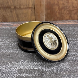 Puede incluir: Una caja decorativa redonda negra y dorada con tapa a juego. La tapa presenta un marco negro con un marco interior dorado y una imagen central de un querubín tocando el arpa. La caja está abierta, revelando un interior dorado.