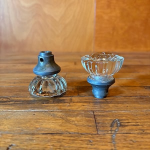 以下が含まれることがあります： 透明で溝のあるファセットカットが施されたガラス製のキャビネットノブ2個。各ノブは円形の透明なガラスのトップと、取り付け用の小さな穴が開いたダークグレーの金属製ベースで構成されており、ヴィンテージ風のスタイルを特徴としています。
