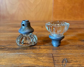 May include: Two glass cabinet knobs with a clear, fluted, and faceted design. Each knob features a circular, transparent glass top and a metallic, dark grey base with a small hole on the side for attachment, showcasing a vintage-inspired aesthetic.