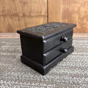 May include: A small, black wooden jewelry box with two drawers and a decorative carved top. The box has a rectangular shape and sits on a small base. The drawers have round knobs.