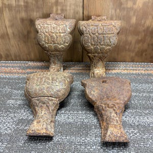 May include: Four antique cast iron furniture feet with a weathered, rusty finish. The feet have a decorative, ornate design with the words "ROOTS" and "OAKS" embossed on them. The feet are likely from a vintage piece of furniture.