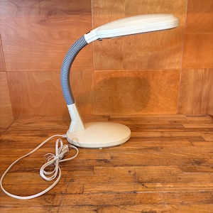 May include: A beige desk lamp featuring a flexible, ribbed grey neck and a wide, oval-shaped light head. The lamp has a sturdy, circular base and includes a white power cord with a standard plug extending from the back of the base unit.