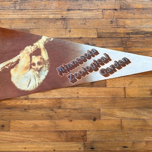 May include: A vintage pennant featuring a brown and white slow loris hanging from a branch. The pennant has the text "Minnesota Zoological Garden" in brown letters. The pennant has a blue border and is on a wooden surface.