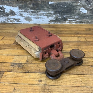 May include: An antique, rusty red metal and wood pulley system. The system includes a rectangular metal piece with bolts, attached to a wooden base, and a separate pulley wheel assembly. The items are on a wooden surface.