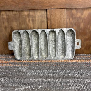 May include: A vintage cast iron cornbread mold in the shape of six ears of corn. The mold is a dull silver color and has handles on each end. The mold is sitting on a gray and orange striped surface.