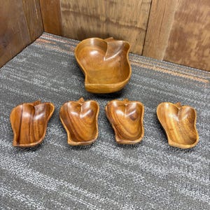 May include: A set of wooden serving bowls and dishes. The large bowl is apple-shaped, and the smaller dishes are also apple-shaped. The wood has a warm, brown tone with visible grain patterns. The set is arranged on a gray surface.