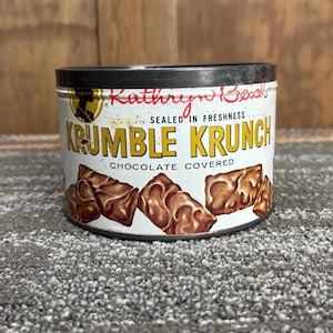 May include: A vintage metal tin of "Krumble Krunch" chocolate-covered biscuits. The tin is white with gold and brown text and illustrations. The top and rim are black. The text "Kathryn Beich" is written in red script.