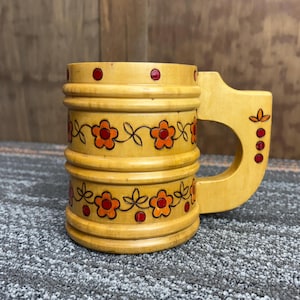 May include: Wooden mug with a handle, featuring a floral design in orange and black. The mug has a light brown color with red accents. The handle is integrated into the design, and the mug has a cylindrical shape.