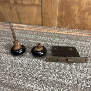 May include: A collection of vintage door hardware, featuring two black ceramic door knobs and a metal door latch. The knobs have a glossy finish and are attached to aged metal stems. The latch is rectangular with a sliding bolt.