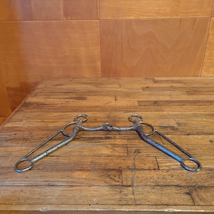 May include: A metal horse bit featuring a jointed mouthpiece connected to two long, angled shanks with integrated rings at the top and bottom. The hardware displays a weathered, textured finish with visible signs of oxidation and metallic wear.