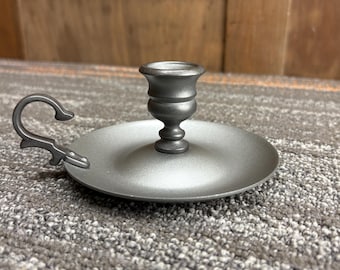 Puede incluir: Un candelero de color peltre con una base circular y una copa central para sostener una vela. Cuenta con un asa curvada en el lateral. El candelero es de metal y tiene un acabado mate. El diseño es sencillo y elegante.