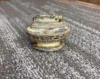 May include: An antique, ornate lighter with a cream and silver finish. The lighter has a rounded body with decorative detailing and a hinged top. The base is also decorated with a repeating pattern. The lighter appears to be made of metal.