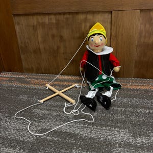 May include: A wooden marionette doll of a boy with a yellow hat, red shirt, black vest, and green polka dot pants. The doll has a white collar and black shoes. The strings are attached to a wooden control.