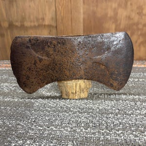 May include: A vintage double-bit axe head with a weathered, rusty appearance. The axe head is mounted on a small, light-colored wooden base. The axe has two blades and is designed for chopping wood.
