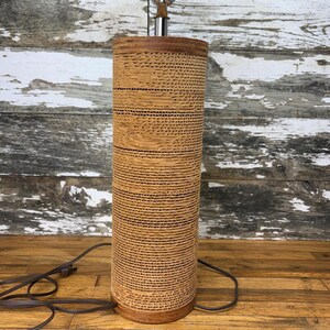 May include: A cylindrical table lamp featuring a textured, layered cardboard shade in a light tan colour. The lamp has a smooth wooden base and a metallic top fitting, with a dark brown electrical cord extending from the bottom of the unit.