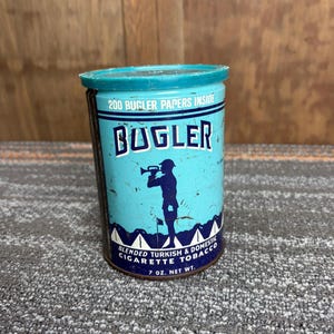 May include: Vintage Bugler cigarette tobacco tin. The cylindrical tin is light blue with a dark blue top. The tin features the word "BUGLER" in white, a silhouette of a bugler, and the text "BLENDED TURKISH & DOMESTIC CIGARETTE TOBACCO".