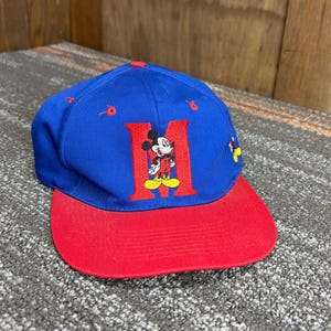 May include: A blue baseball cap with a red brim and red eyelets. The front features a large red letter M with an embroidered Mickey Mouse character standing in front of it. A small yellow and red Mickey Mouse icon is visible on the side panel.
