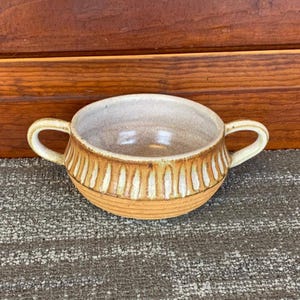 May include: A handmade ceramic soup bowl with two handles. The bowl is tan with vertical white stripes around the middle and a light brown interior. The bowl sits on a gray and brown surface.