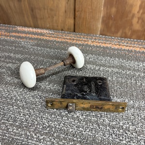 Puede incluir: Juego de herrajes de puerta antiguos que presenta un pomo de porcelana blanca con una varilla de metal oxidado, un pestillo de metal negro y una placa de cierre de latón. El juego es probablemente de principios del siglo XX.