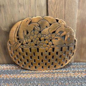May include: A copper-colored metal wall hanging in the shape of a basket filled with fruit. The basket has a rectangular cut-out design. The fruit includes grapes and other shapes.