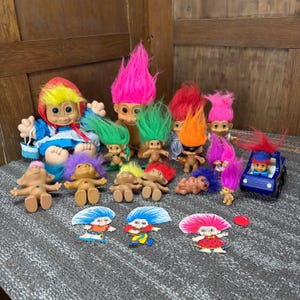 May include: A collection of various troll dolls featuring vibrant hair in shades of pink, green, orange, yellow, and purple, alongside a blue toy car, a larger doll in a blue dress, and three flat paper cutouts of troll characters.
