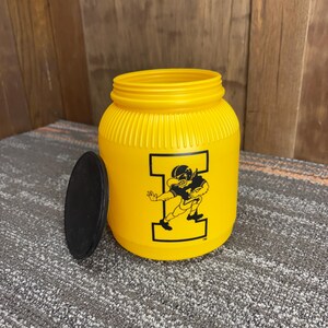 May include: Yellow plastic jar with a black lid. The jar features a black graphic of a football player inside a large letter "I". The jar has a ribbed texture and a wide opening.