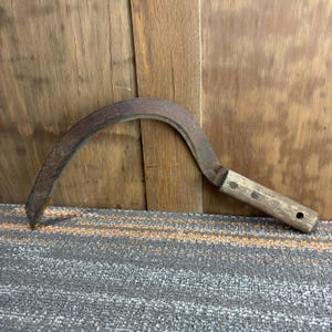 Puede incluir: Una hoz antigua con una hoja curva de metal oxidado y un mango de madera. El mango está sujeto con pequeños remaches metálicos. La herramienta, diseñada para la cosecha, tiene un aspecto clásico y rústico.