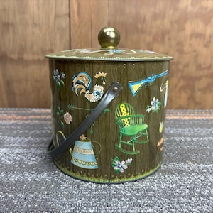 Puede incluir: Cubo de metal verde oliva vintage con tapa y asa negra. El cubo está decorado con ilustraciones coloridas de un gallo, una mecedora, flores y una tetera. La tapa tiene un pomo dorado.