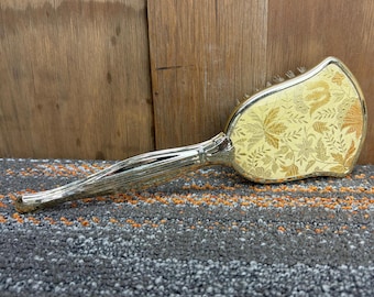 Puede incluir: Un cepillo para el cabello vintage con un diseño floral dorado sobre un fondo crema. El mango es plateado y tiene una textura acanalada. El cepillo se muestra sobre un fondo de madera.