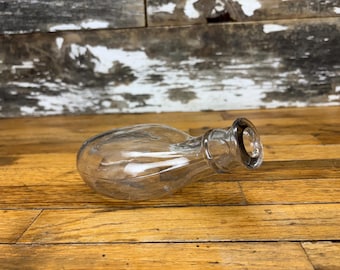 May include: A clear glass bottle with a rounded body and a flared neck. The bottle is lying on its side on a wooden surface. The background is a weathered wooden wall.