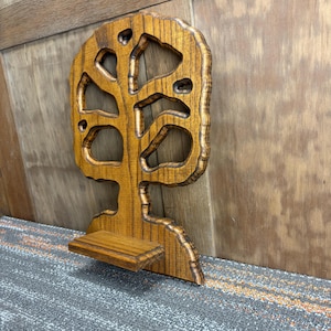 May include: A wooden tree-shaped wall shelf with a natural wood grain finish. The shelf has a small rectangular platform at the bottom and cut-out shapes within the tree design. The shelf is designed to be mounted on a wall.