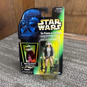 May include: A Star Wars Rebel Fleet Trooper action figure in its original packaging. The card features Darth Vader's silhouette, the Star Wars logo, and the text "The Power of the Force". The figure is holding a blaster pistol and rifle.
