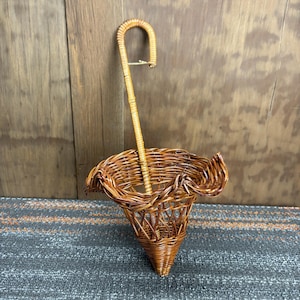 May include: A brown wicker umbrella stand with a curved handle. The stand has a cone shape and a ruffled top edge. The wicker is a warm brown colour, and the stand is designed to hold umbrellas. The background is a wooden wall.