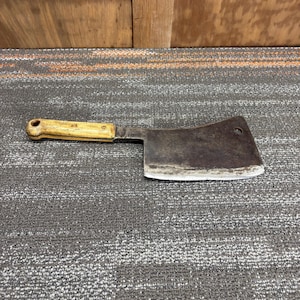 May include: A vintage meat cleaver with a worn, dark metal blade and a light-colored wooden handle. The cleaver has a rectangular blade with a slightly curved cutting edge and a hole near the top. The handle is attached with rivets.