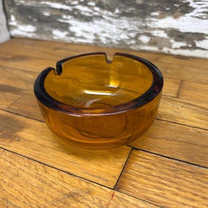 May include: Amber glass ashtray with a round shape and a small indentation for cigarettes. The ashtray is sitting on a wooden surface.