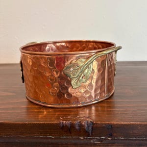 Puede incluir: Un cuenco de metal martillado de color cobre con detalles de hojas de latón. El cuenco tiene forma redondeada y superficie texturizada. Las hojas de latón están unidas a los lados, añadiendo un toque decorativo. Una pieza única para la decoración del hogar.