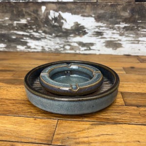 May include: A blue ceramic ashtray with a smaller inner bowl, set on a matching blue ceramic tray. The tray has a textured surface.