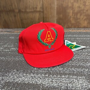May include: A red baseball cap with a flat brim. The cap features a green laurel wreath design with a yellow letter "A" in the center. The word "AUSTRALIA" is embroidered in blue below the design.