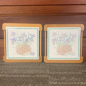 Puede incluir: Dos posavasos de cerámica con marco de madera con un diseño floral de iris morados en una cesta marrón. Los posavasos tienen un borde verde y blanco.