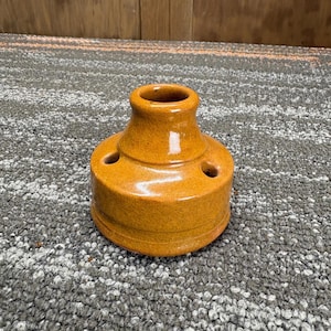 May include: A ceramic, mustard-colored candle holder with a round base and a flared top. The candle holder has three small holes around the base and a single hole at the top. The surface has a crackled texture.