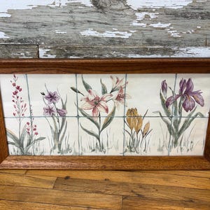 May include: A framed floral tile art piece. The artwork features a variety of flowers, including lilies and irises, in shades of pink, purple, and yellow. The tiles are set in a wooden frame, creating a decorative wall hanging.
