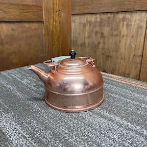 May include: A copper kettle with a rounded body, spout, and a lid with a black knob. The handle is made of wood and metal. The kettle is sitting on a gray surface.
