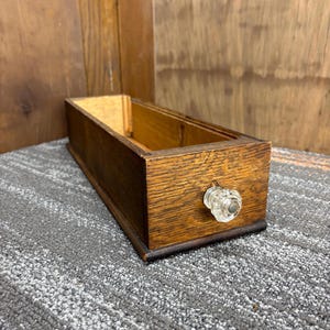 May include: A rectangular wooden drawer with a clear glass knob. The drawer is made of light brown wood with visible grain and a dark brown base. The drawer is empty and sits on a grey carpet.