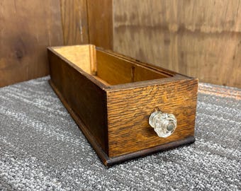Puede incluir: Cajón rectangular de madera con un pomo de cristal transparente. El cajón es de madera marrón claro con vetas visibles y una base marrón oscuro. El cajón está vacío y sobre una alfombra gris.