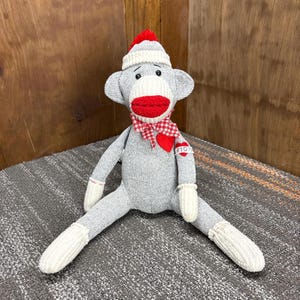 Puede incluir: Un muñeco de mono de calcetín gris con un gorro rojo y blanco, labios rojos y una bufanda a cuadros rojos y blancos. El muñeco tiene guantes y calcetines blancos. Un corazón rojo con la palabra "Mamá" está unido a la bufanda.