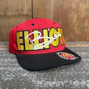 Puede incluir: Gorra de béisbol roja con visera negra. La gorra presenta el nombre "ELLIOTT" en letras amarillas con una firma blanca. Un logotipo de McDonald's Racing Team está en la visera.